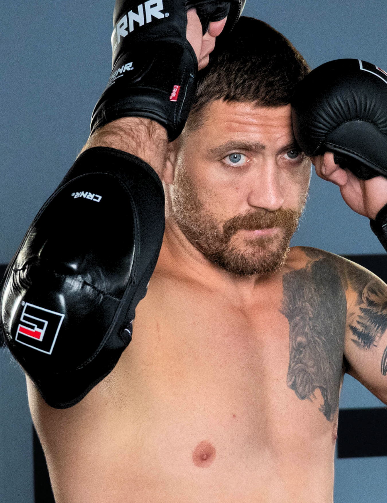 Close-up portrait of a UFC Fighter and Combat Corner Pro Team Member Gerald Meerschaert shirtless with blue eyes and a lion tattoo, holding a high-guard defensive position while wearing Combat Corner’s black Prime MMA Sparring Gloves and Premium Elbow Pads.