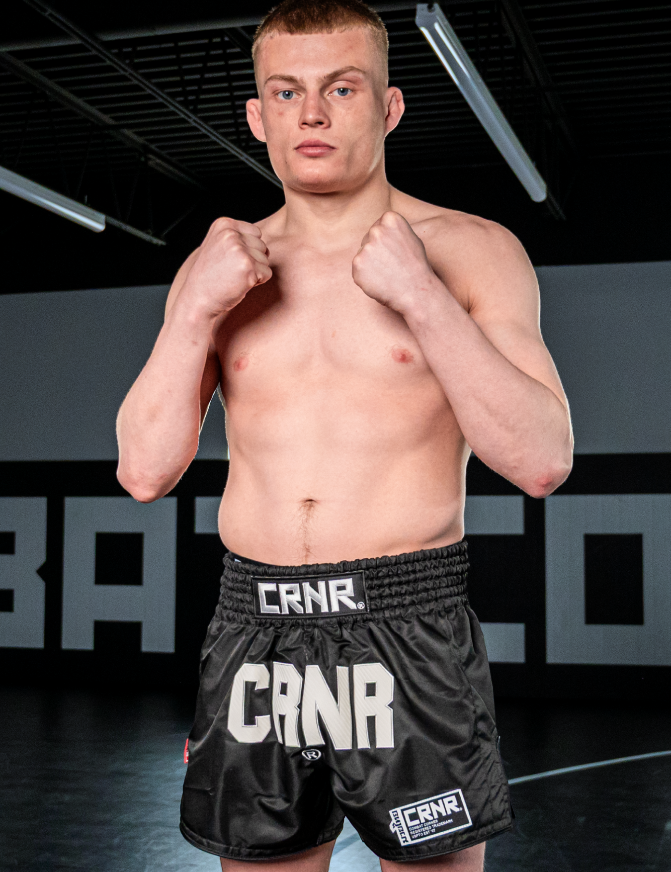 Full-body studio portrait of a shirtless pro fighter and Combat Corner Pro Team Member Jeremy Stroble standing in a guard stance with raised fists, wearing Combat Corner’s black HMIT Supply Muay Thai Shorts featuring prominent white "CRNR" branding logos. 