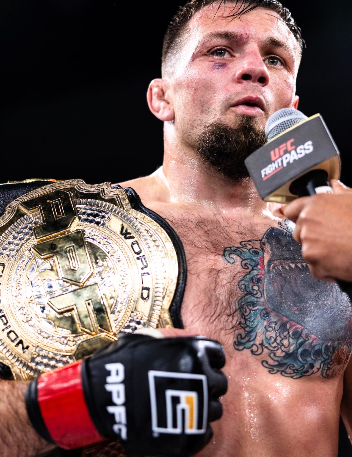 Close-up of a victorious Lightweight APFC Champion and Combat Corner Pro Team Member Mark Choinski with a shark chest tattoo speaking into a UFC Fight Pass microphone, wearing black Combat Corner APFC MMA Fight Gloves with red wrist cuffs while draped in a gold championship belt. 