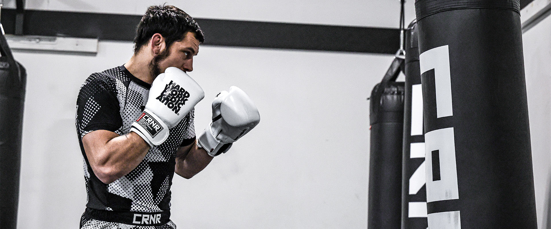 Professional fighter Nick Klein in a boxing stance facing a heavy bag in a gym, wearing Combat Corner’s HWD Boxing Gloves and Camcow Rash Guards and Combat Trainers Fight Shorts. 