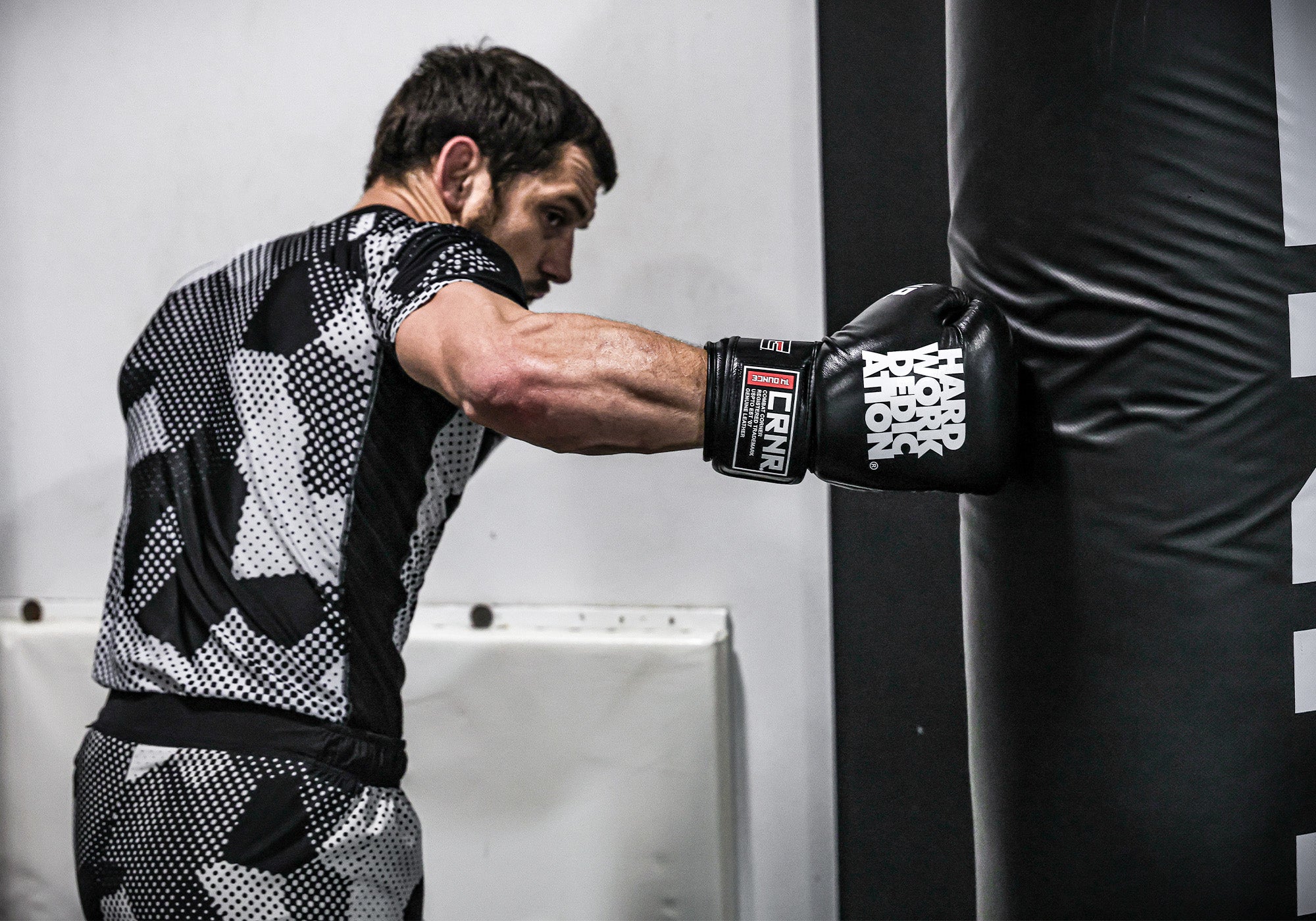 Wide portrait of professional fighter Nick Klein striking a heavy bag while wearing Combat Corner’s black HWD Boxing Gloves and Combat Corner’s Camcow Rash Guard, a black and white patterned compression shirt. 