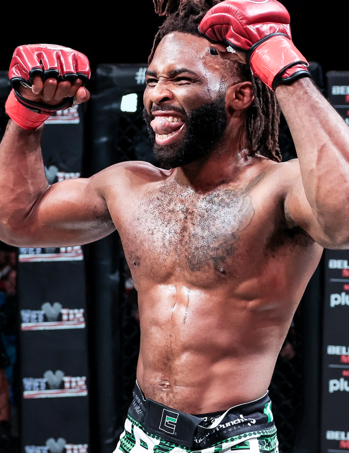 Close-up of a victorious Bellator Bantamweight  Champion and Combat Corner Pro Team Member Raufeon Stots with dreadlocks and a beard as he flexes both arms inside the cage, sticking out his tongue while wearing red Combat Corner fight gloves and patterned green fight shorts. 