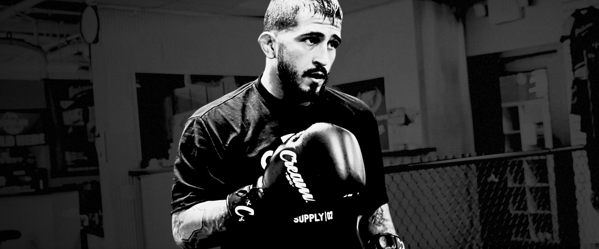 A black and white panoramic portrait of pro fighter and Combat Corner Pro Team Member Sergio Pettis looking focused during training, wearing a black Combat Corner t-shirt and Cream Boxing Gloves.