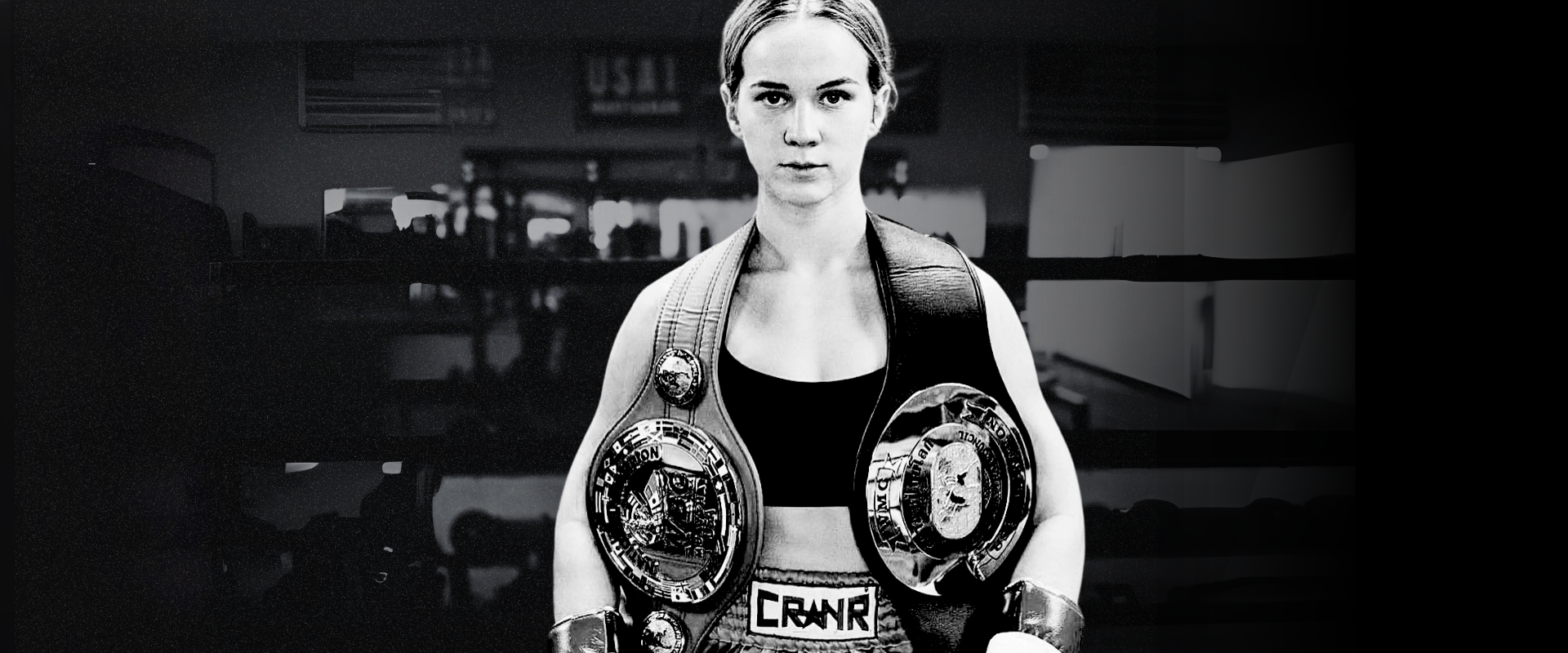 High-contrast black and white portrait of professional fighter Tierra Brandt looking forward with two championship belts draped over her shoulders, wearing Combat Corner Muay Thai Shorts with a prominent "CRNR" waistband logo.