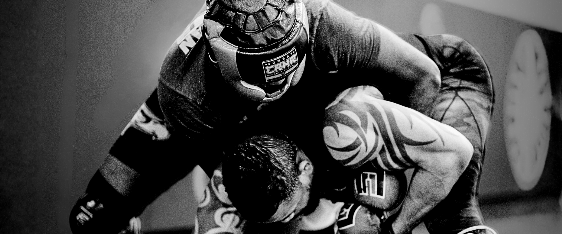 A black and white close-up shot of two fighters grappling on a mat, with the top fighter wearing Combat Corner’s HMIT Headgear featuring a prominent "CRNR" forehead label. 
