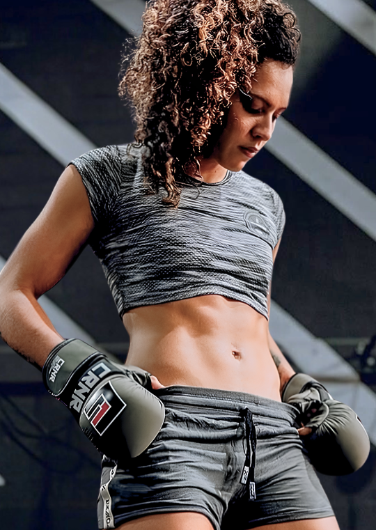 Low-angle portrait of a female martial artist with curly hair looking down, wearing Combat Corner’s OD Green MMA Spar Gloves, a space-dye grey crop top, and athletic training shorts. 