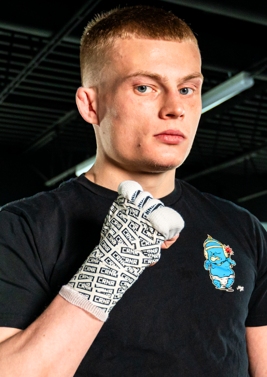 A portrait of a young male fighter looking at the camera with his fist raised, showing off Combat Corner Hi-Tack Combat Tape covered in a repeating black Combat Corner "CRNR" logo pattern. 