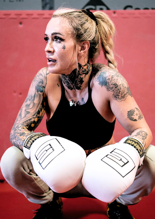 Candid portrait of a heavily tattooed female fighter crouching in a gym, wearing a black sports bra, grey sweatpants, and white Combat Corner HMIT Boxing Gloves with gold wrist labels. 