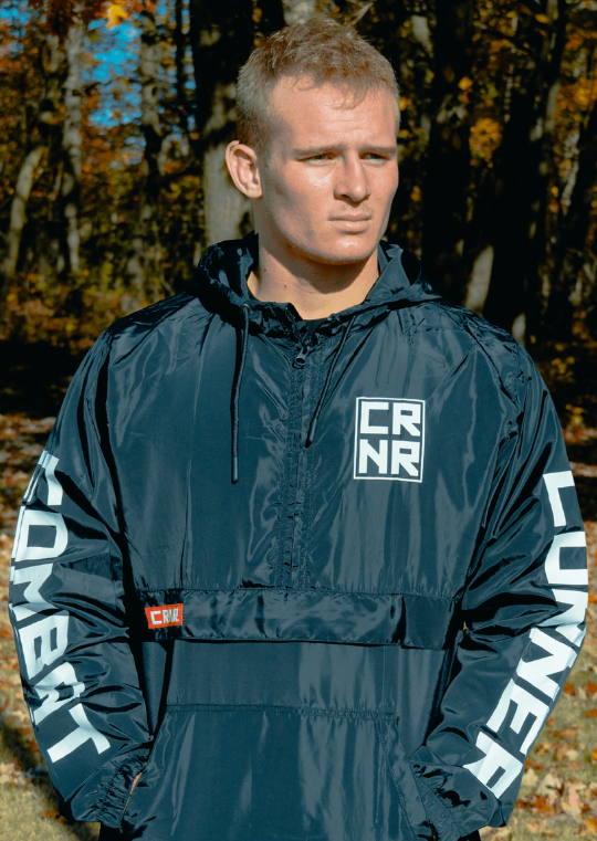 Medium portrait of a male athlete outdoors wearing a black Combat Corner Windbreaker, which is a quarter-zip windbreaker anorak jacket with bold white text on the sleeves.
