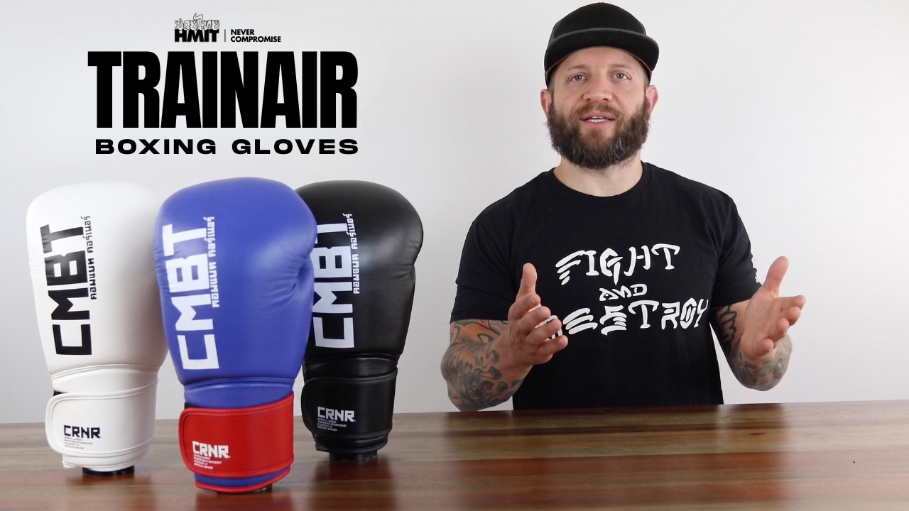 A YouTube thumbnail image featuring pro fighter Dan LaSavage giving a product walkthrough of HMIT TrainAir Boxing Gloves and gesturing toward an array of colorful Combat Corner HMIT TrainAir Boxing Gloves lined up on a wooden table. 