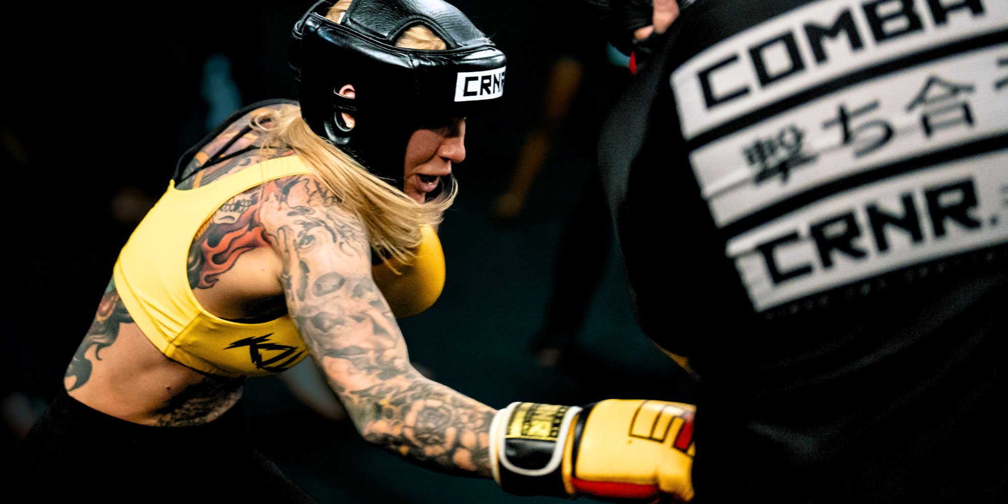 An action-packed shot of a tattooed female fighter throwing a body shot during a sparring session, wearing yellow Combat Corner HMIT Boxing Gloves and Combat Corner black Competition Headgear, while her coach wears a Combat Corner JPN Rash Guard. 