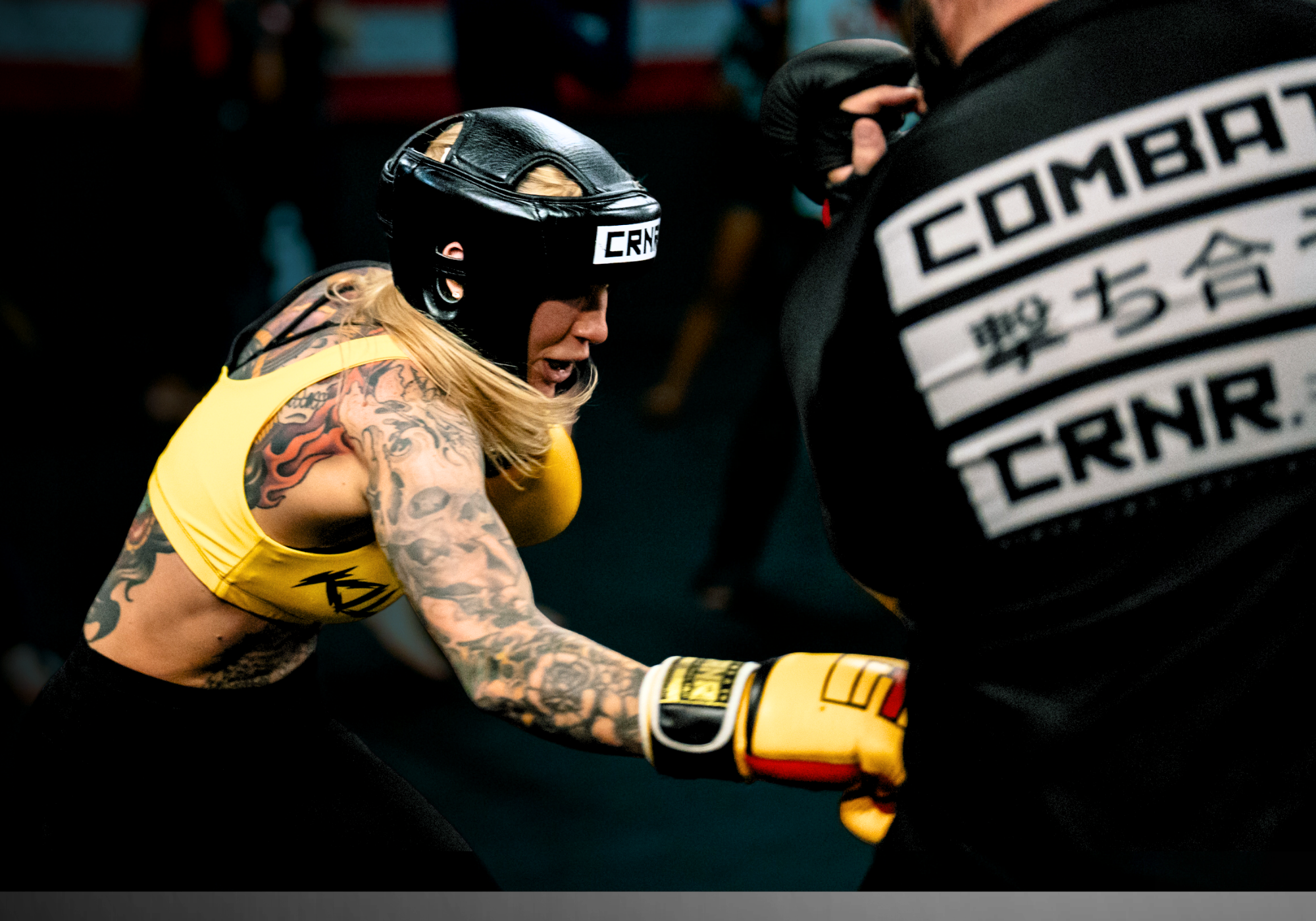 An action-packed shot of a tattooed female fighter throwing a body shot during a sparring session, wearing yellow Combat Corner HMIT Boxing Gloves and Combat Corner black Competition Headgear, while her coach wears a Combat Corner JPN Rash Guard. 