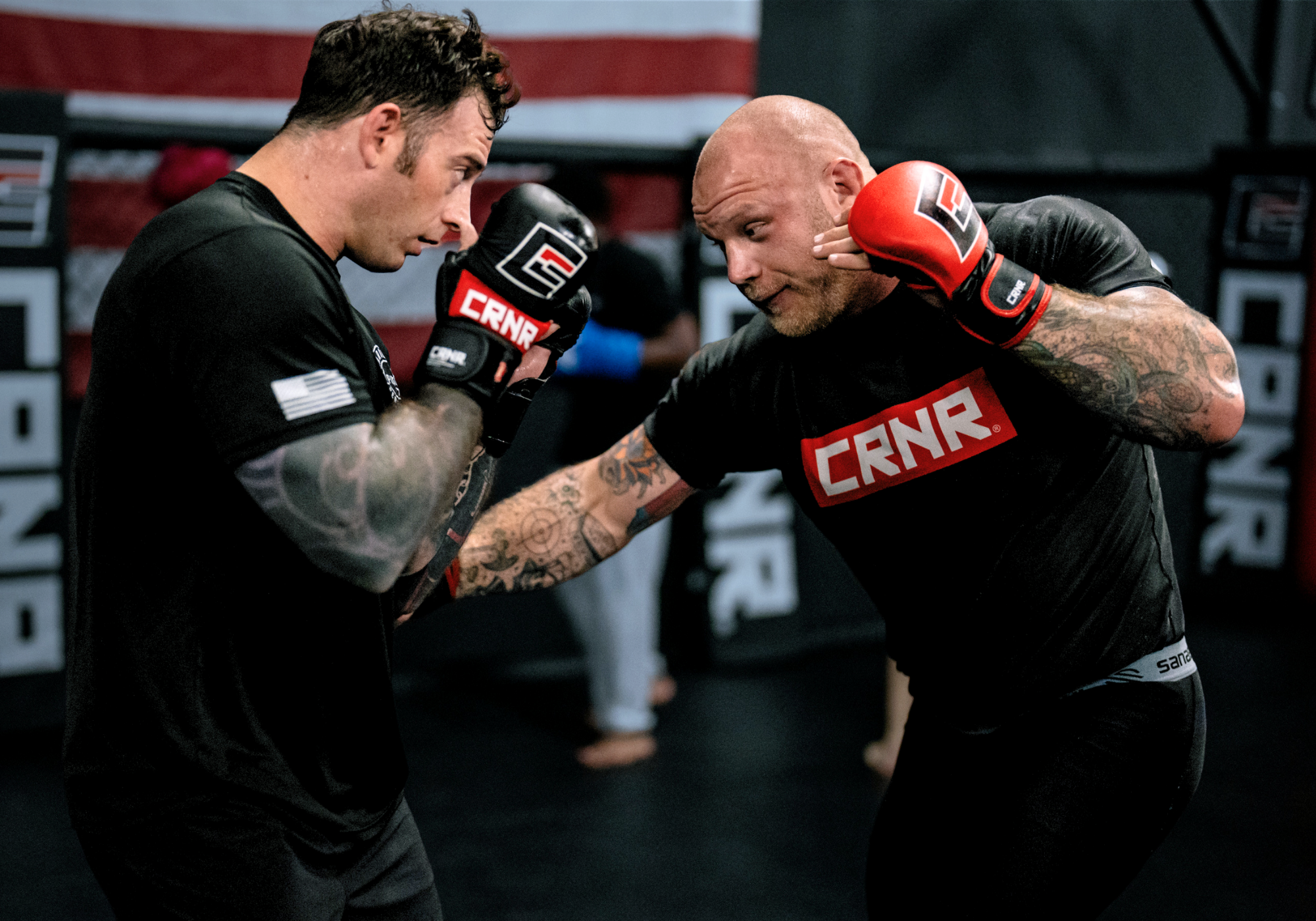 An action shot of two tattooed male fighters sparring in a gym, with one throwing a punch wearing red Combat Corner MMA gloves, while his partner blocks using black Combat Corner sparring gloves.