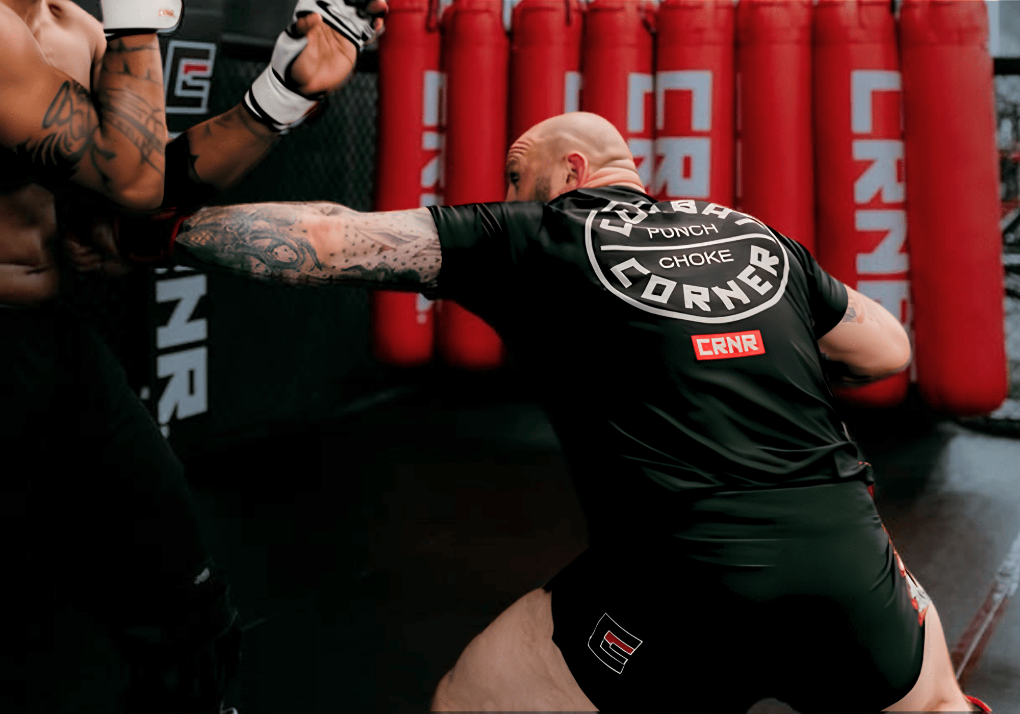 
An action-packed shot of a tattooed male fighter throwing a punch during a training session, wearing a black Combat Corner Rash Guard and Fight Shorts, with red Combat Corner heavy bags in the background. 
