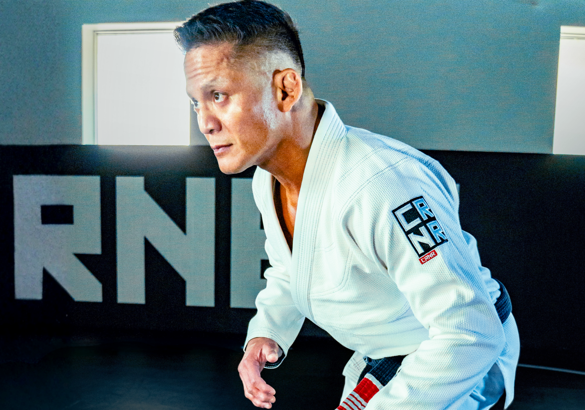 Side profile of a BJJ World Champion Mark Vives in a combat stance, wearing a white Combat Corner V7 Brazilian Jiu-Jitsu (BJJ) Gi with a black belt, featuring a prominent blue and white "CRNR" logo patch on the left sleeve. 