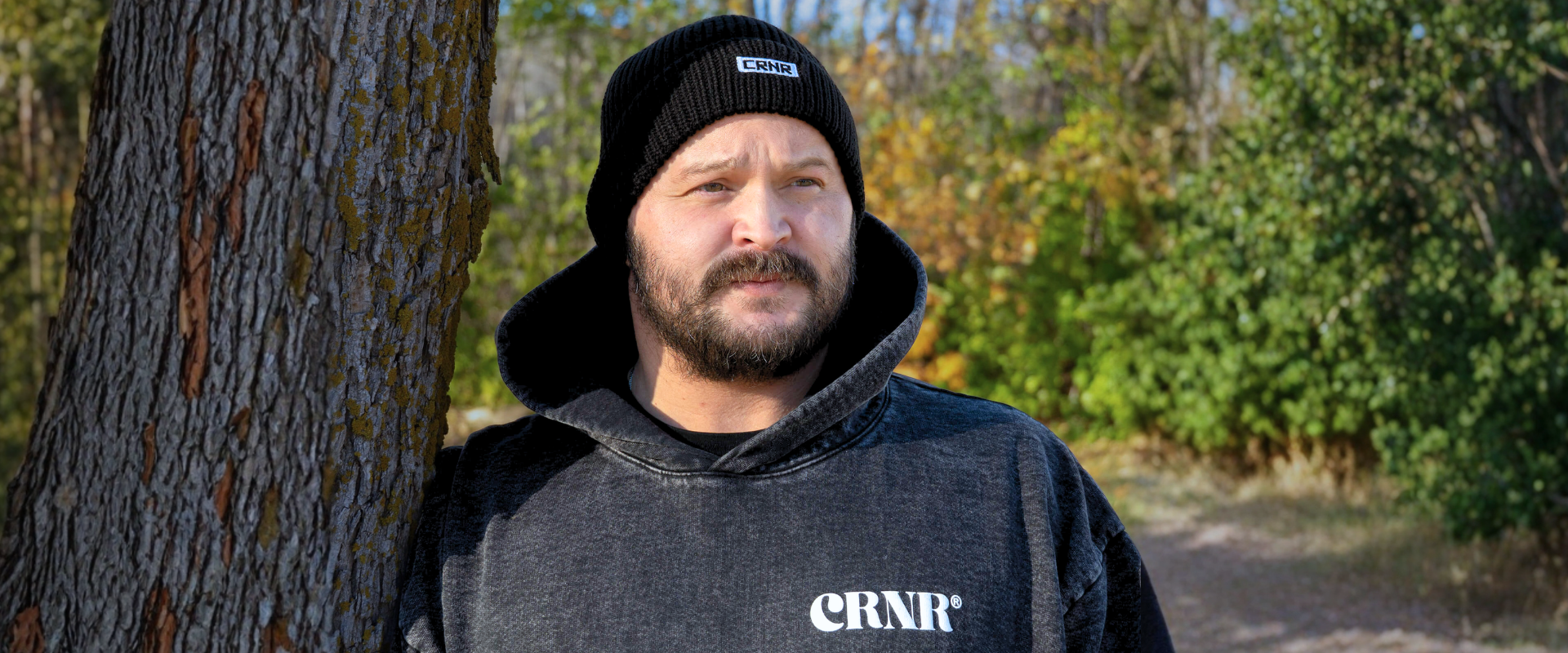 Medium shot of a bearded man leaning against a tree trunk outdoors, wearing Combat Corner’s Relic Hoodie and Trademark Beanie, a black knit cuff beanie with a small "CRNR" logo patch. 