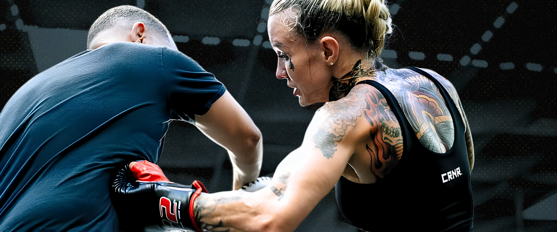 Dynamic action shot of pro female fighter Taylor Starling mid-training session, striking a male sparring partner while wearing a black Combat Corner sports bra featuring a small white "CRNR" logo on the back and Combat Corner’s C2 MMA Training Gloves and HMIT Supply Muay Thai Shorts. 