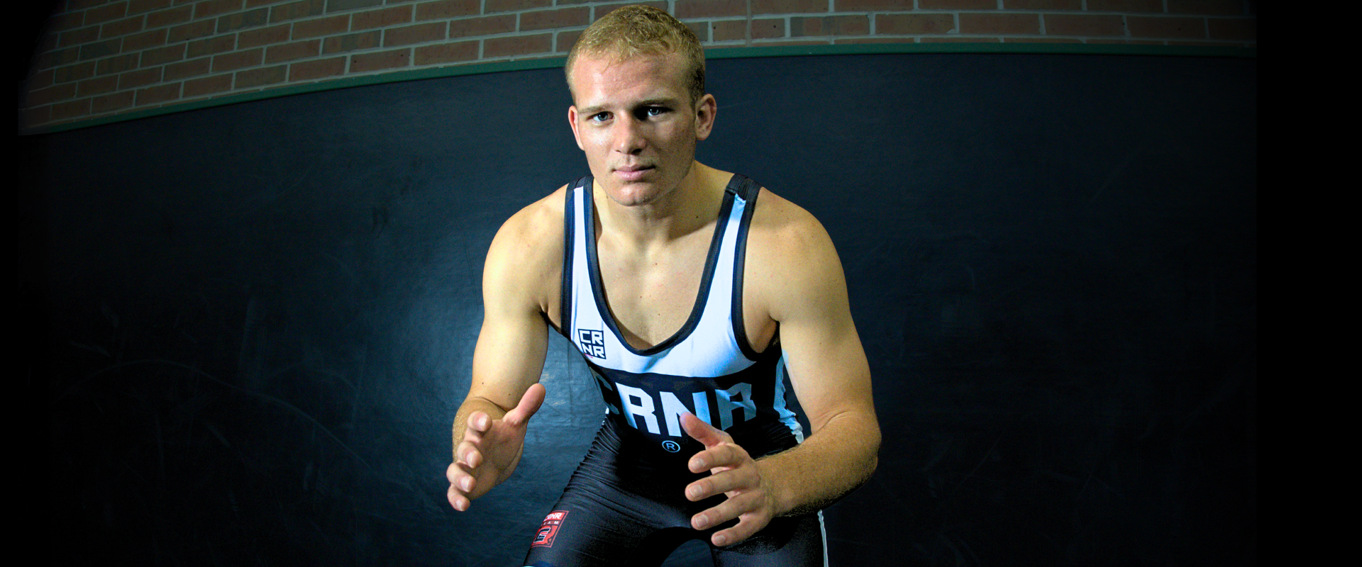 A blonde male wrestler in a low wrestling stance facing the camera, wearing Combat Corner’s Champion Air Wrestling Singlet with a prominent "CRNR" chest graphic. 