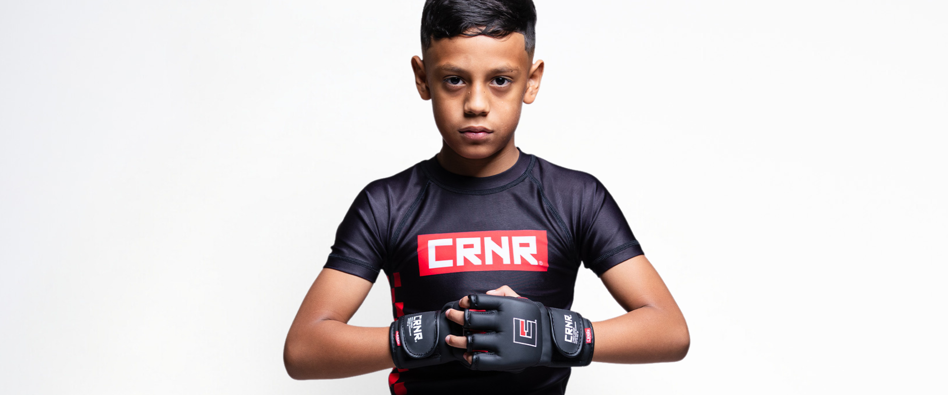 A studio portrait of a serious youth MMA fighter looking forward, wearing a black Combat Corner Kids Rash Guard with a red and white "CRNR" chest logo, clasping his hands together while wearing Combat Corner’s black Kids MMA Spar Gloves. 