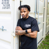 A man with dreadlocks and tattoos on his arms stands with his arms crossed, leaning against a white shipping container. He is wearing a black Combat Corner t-shirt featuring a small, white stylized "CRNR" logo on the left chest.