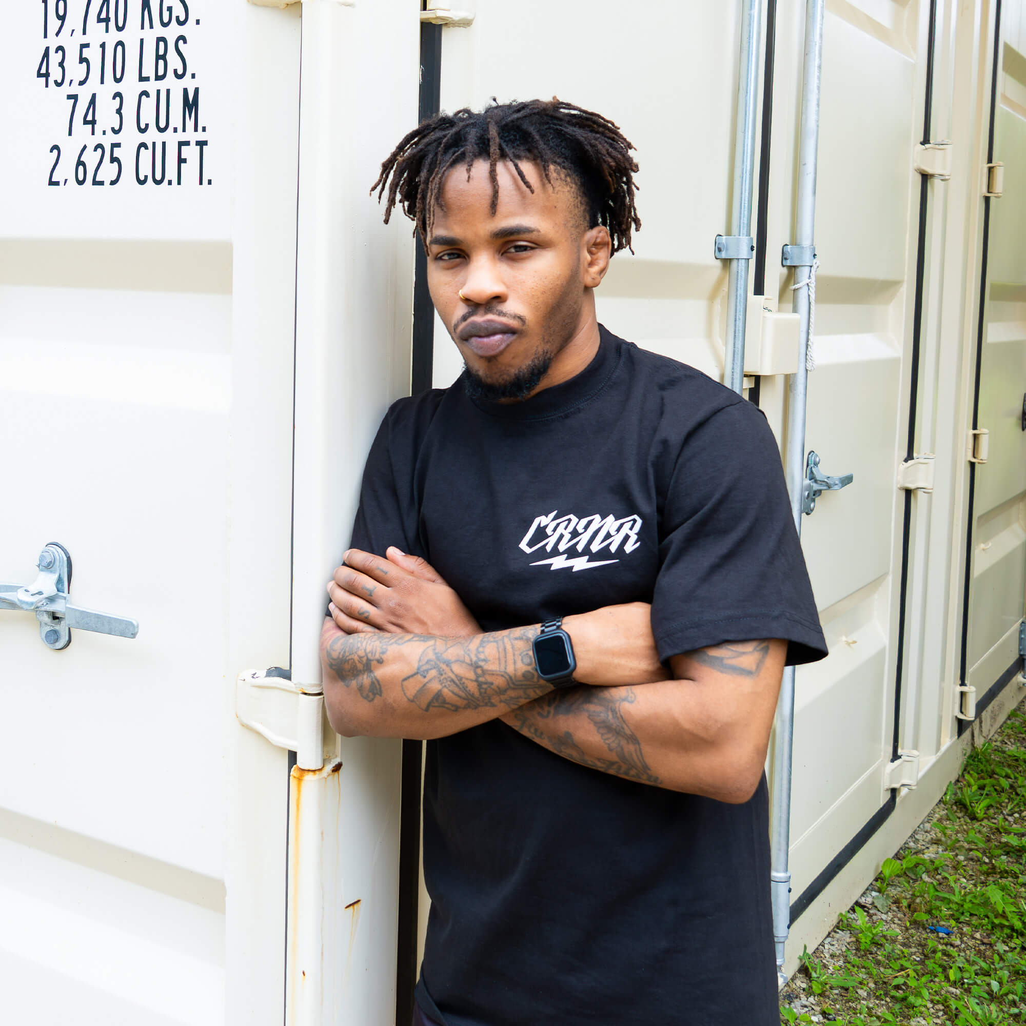 A man with dreadlocks and tattoos on his arms stands with his arms crossed, leaning against a white shipping container. He is wearing a black Combat Corner t-shirt featuring a small, white stylized "CRNR" logo on the left chest.