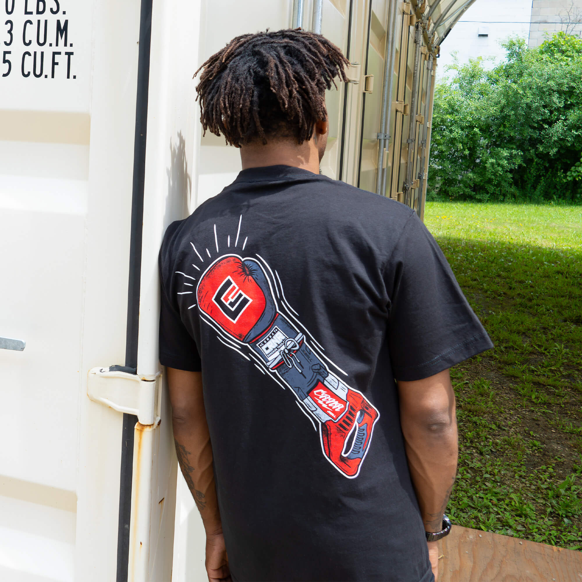 Rear view of a man leaning against a white shipping container, showcasing the back graphic of a black Combat Corner t-shirt. The graphic features a detailed illustration of a red and black boxing glove seamlessly blending into the body of a power tool, with a small "CRNR" logo visible on the design.
