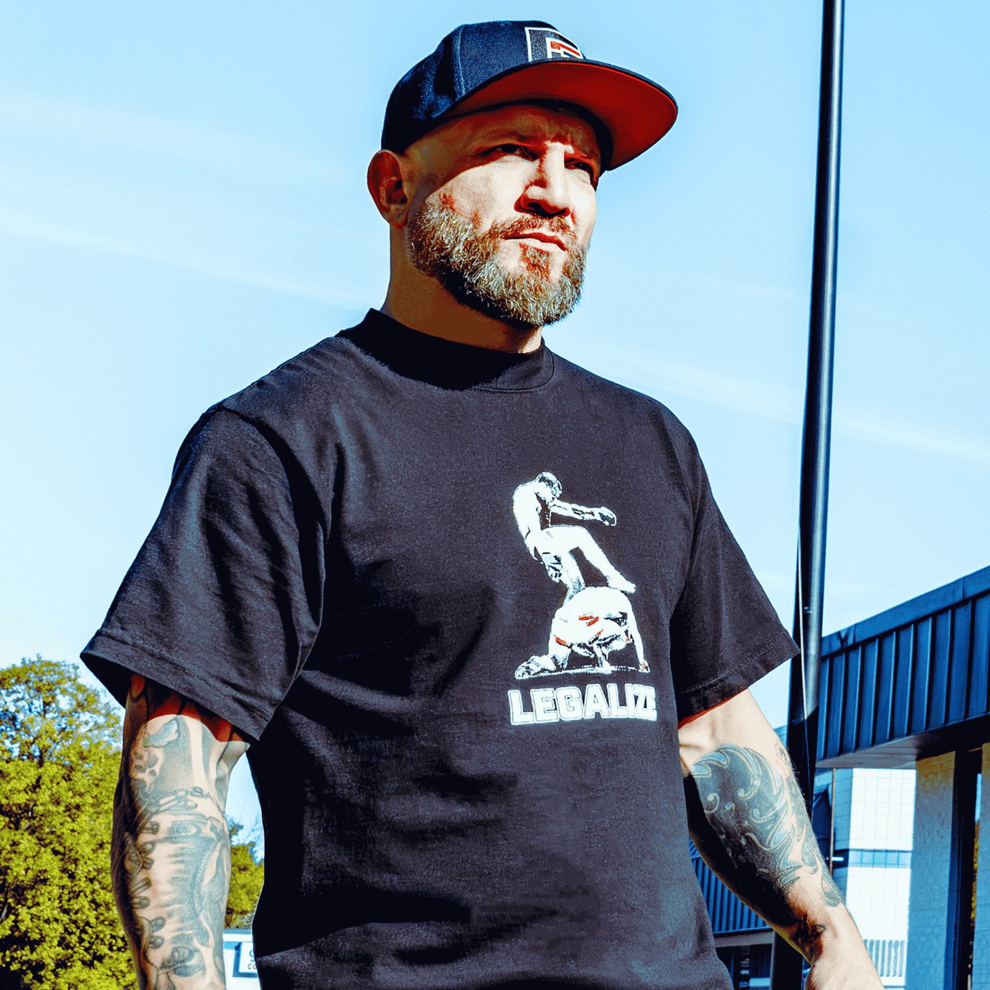 A lifestyle front view photo of a man with arm tattoos wearing the black Combat Corner Legalize Soccer Kicks T-Shirt and a Combat Corner hat standing outside. 