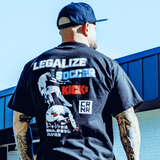 A lifestyle back view photo of a man with arm tattoos wearing the black Combat Corner Legalize Soccer Kicks T-Shirt and a Combat Corner hat standing outside. 