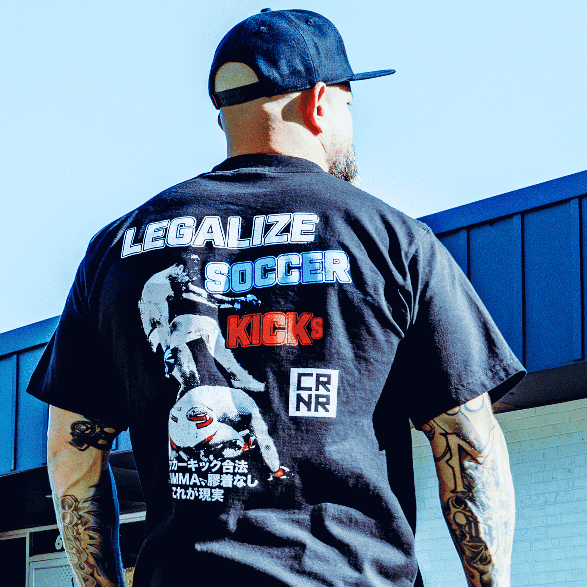 A lifestyle back view photo of a man with arm tattoos wearing the black Combat Corner Legalize Soccer Kicks T-Shirt and a Combat Corner hat standing outside. 
