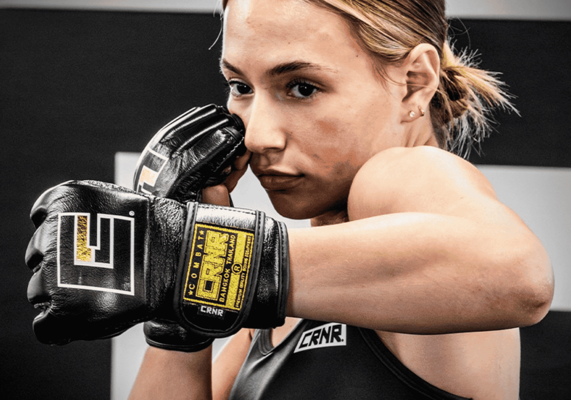A close-up action portrait of a female Muay Thai holding a defensive guard posture while wearing black Combat Corner HMIT MMA Fight Gloves and a branded tank top.