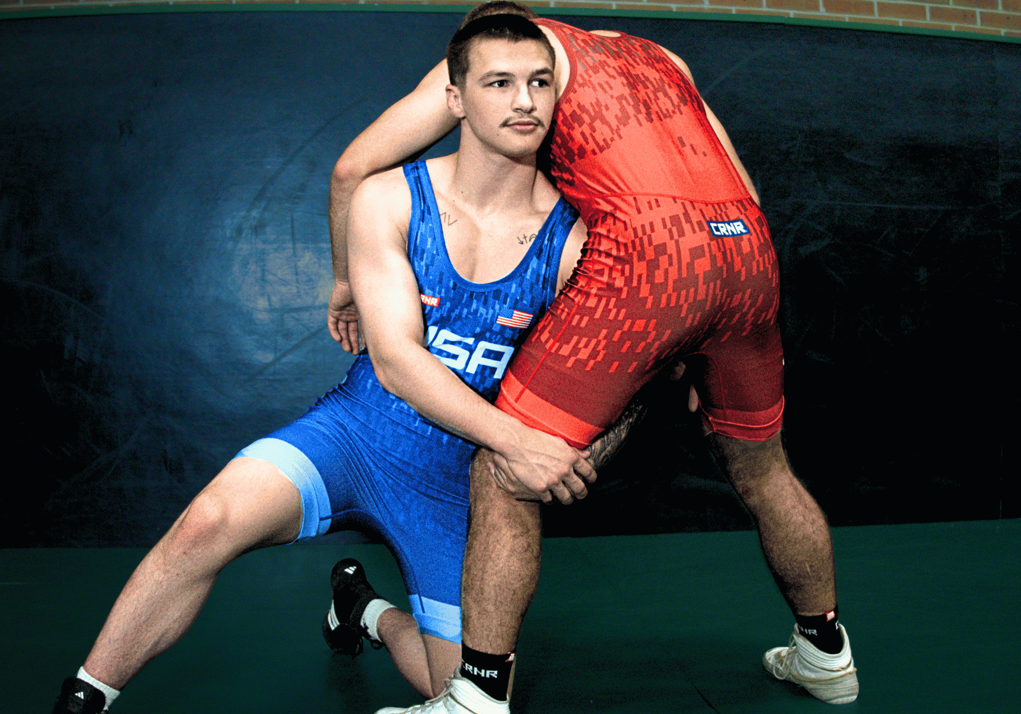An action photograph of two wrestlers training on a mat, with one athlete in a blue Combat Corner USA Freco 25 Air Singlet defending a takedown from an opponent wearing a red Combat Corner USA Freco 25 Air Singlet.