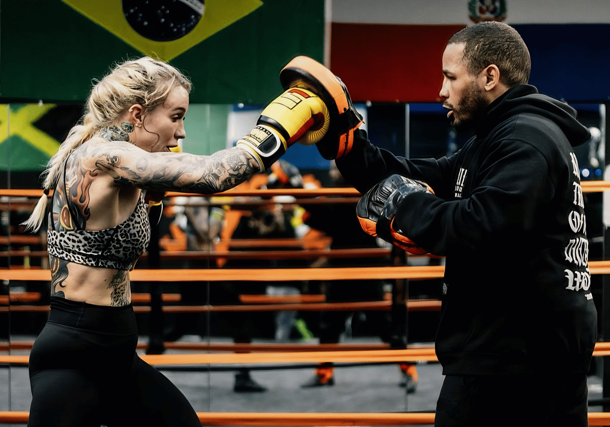 An action shot of a heavily tattooed female fighter striking a focus mitt held by her coach during a boxing training session in a ring, using Combat Corner training gear.