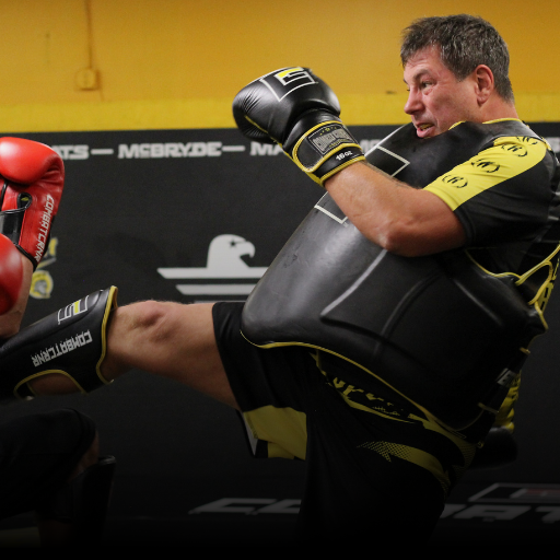 An action shot of legendary coach Duke Roufus training in a gym, wearing black and yellow Combat Corner 16 oz boxing gloves, matching shin guards, and a black protective body shield while throwing a kick.