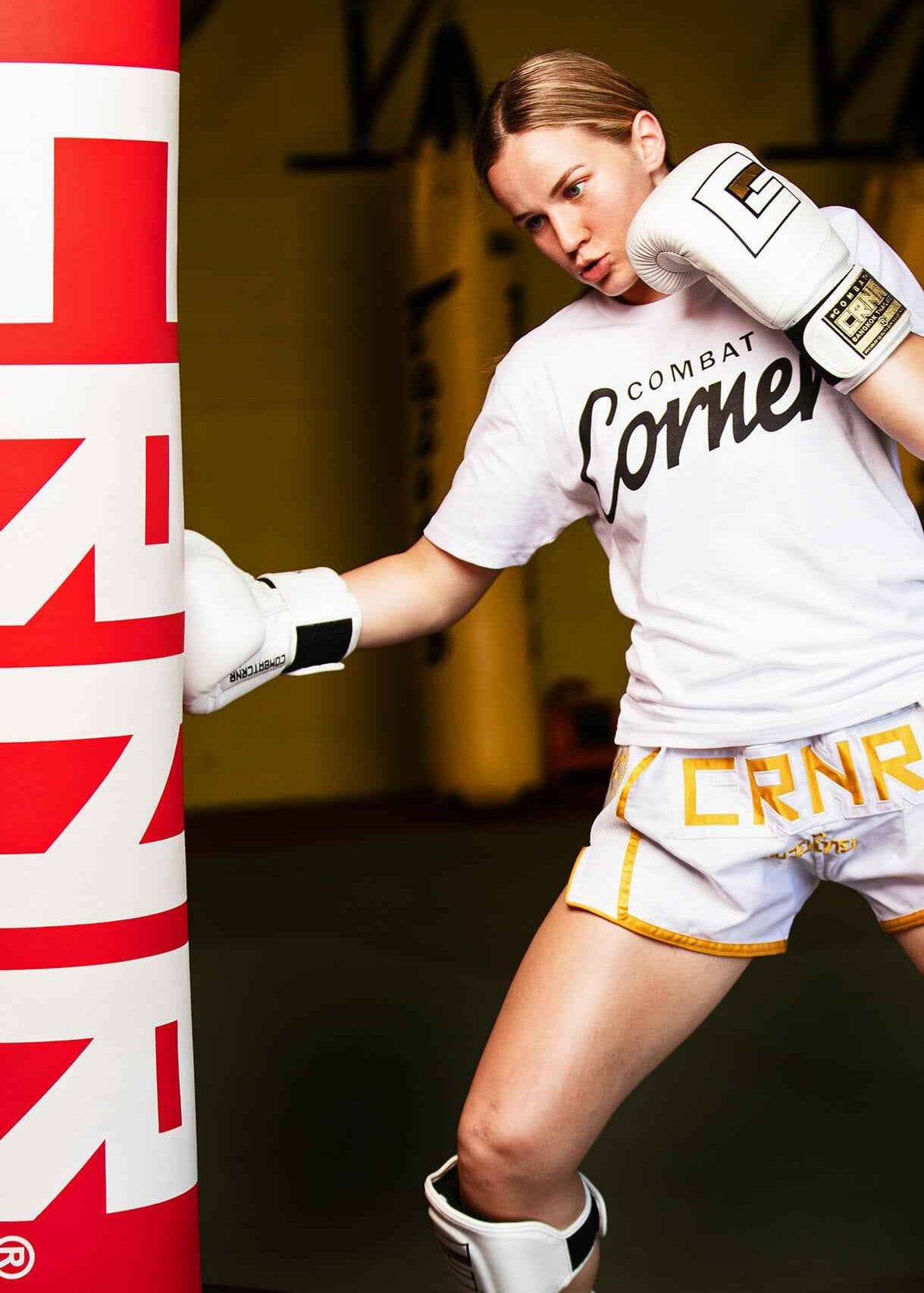 A photo of Muay Thai world champion Tierra Brandt striking a red and white HMIT heavy bag, training with a white Combat Corner t-shirt, white and gold Combat Corner Muay Thai shorts, white HMIT Boxing Gloves, and white HMIT Shin Guards. 