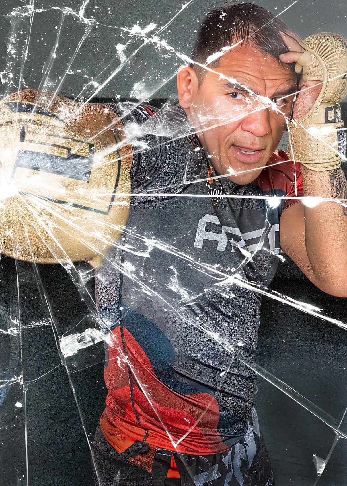 An action shot of an MMA fighter throwing a straight punch while wearing tan Combat Corner HMIT MMA Training Gloves, styled with a dramatic shattered glass visual effect overlay.