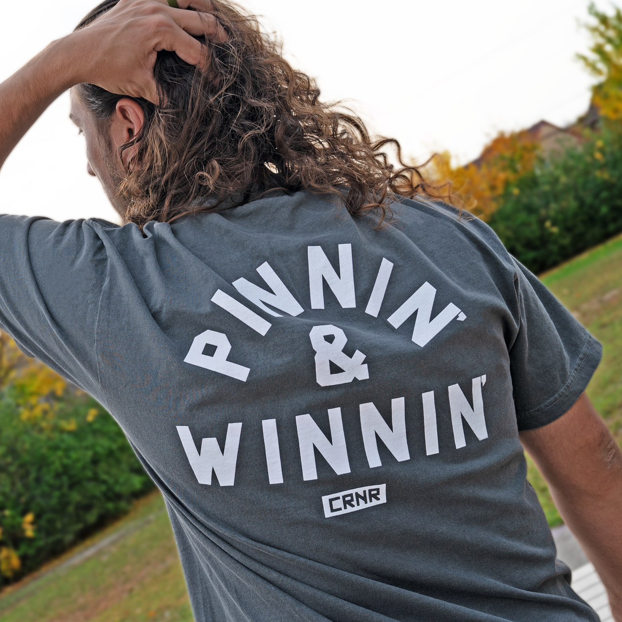 Product photo of Combat Corner’s exclusive Pinnin’ & Winnin’ Relaxed Tee. This photo shows a young man wearing Combat Corner’s Pinnin’ & Winnin’ Faded Navy Relaxed Tee. The photo shows the back of the CRNR tee. The man is outside his local gym after his workout and wrestling training session. This CRNR tee offers the same softness and comfort as your favorite, well-loved vintage tee.