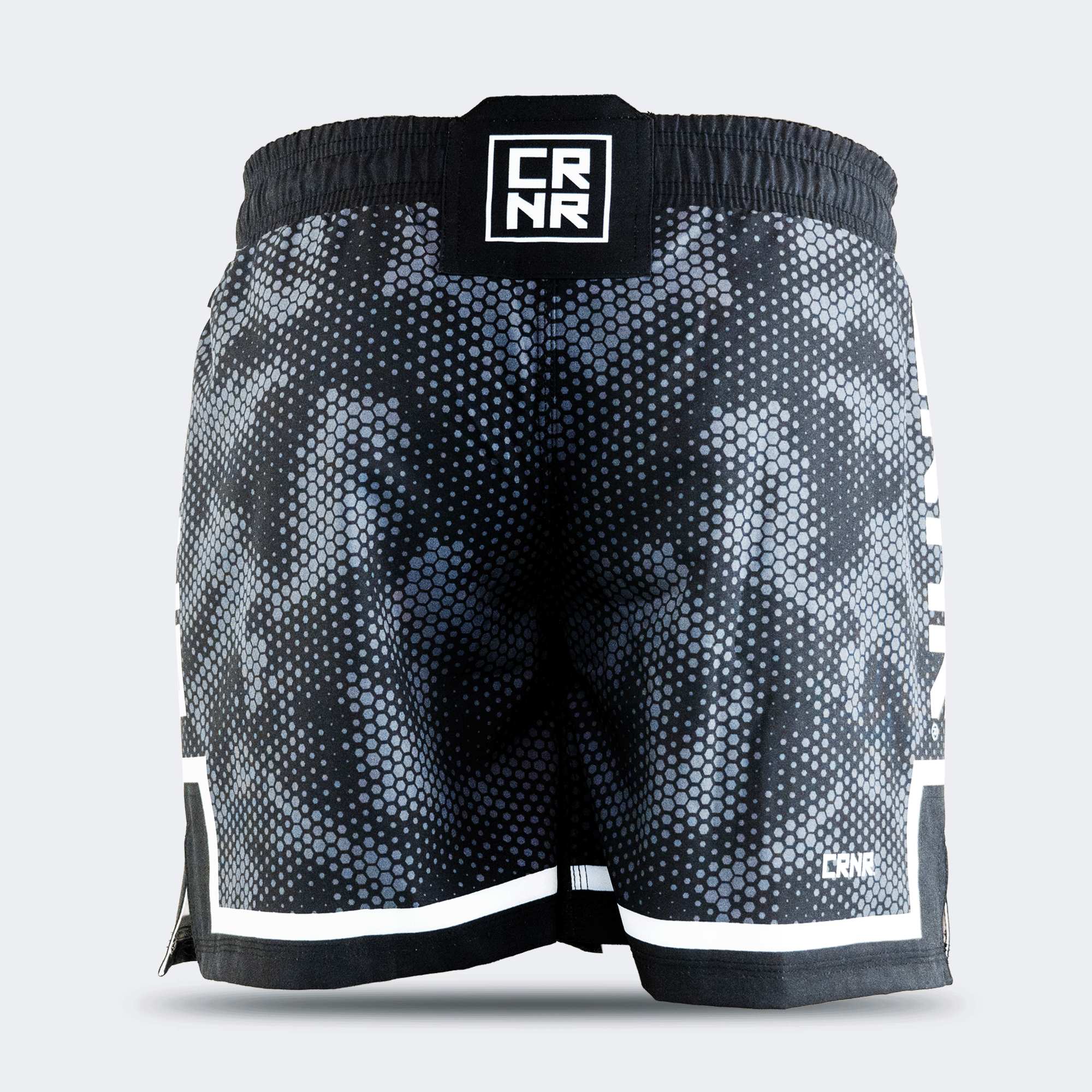 Rear view of Combat Corner CRNR ISO black fight shorts. Displays a centered, square black interlocking "CRNR" logo patch on the gathered elastic waistband. A thin white horizontal stripe runs just above the black bottom hem on both legs, and a small white logo print sits on the lower right leg.