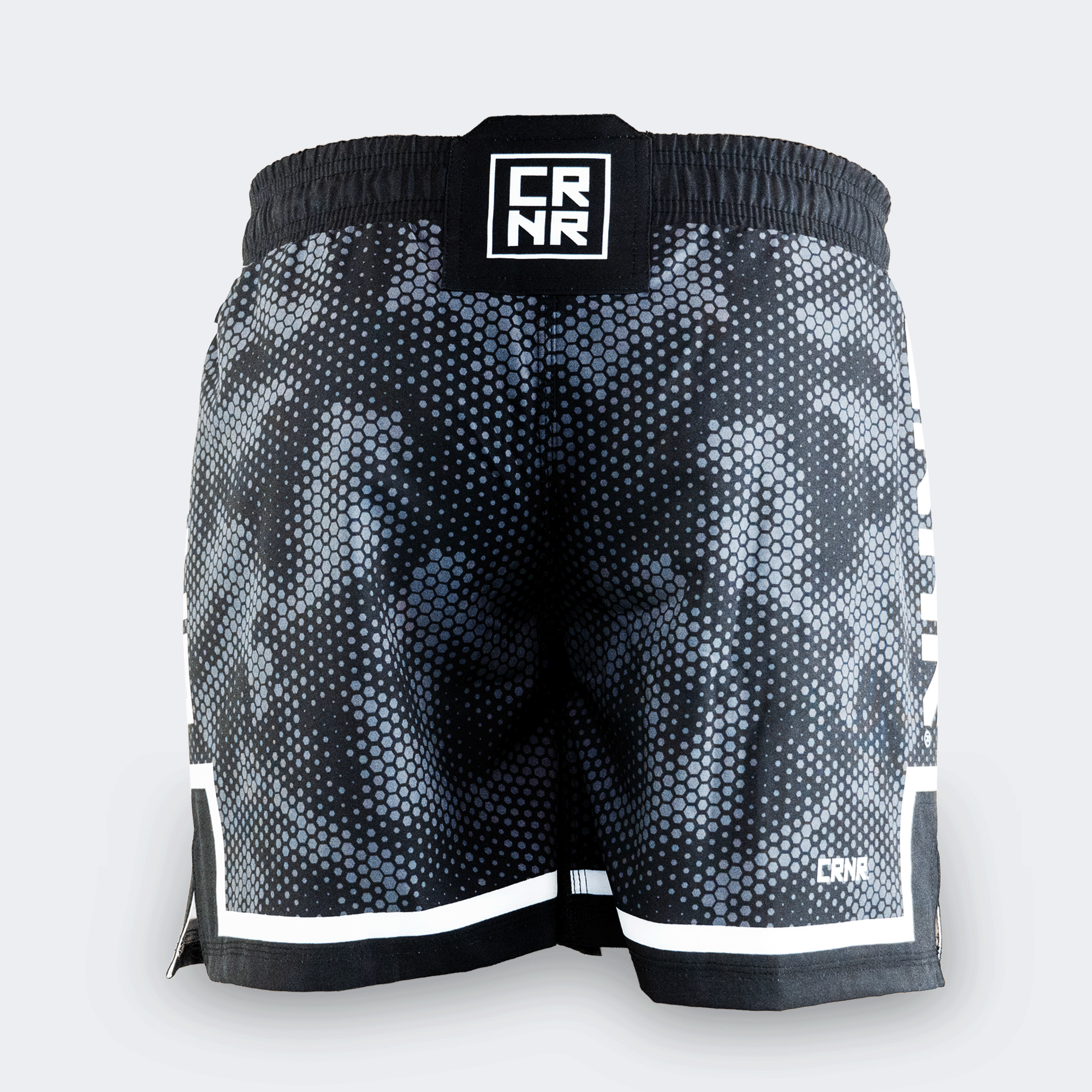 Rear view of Combat Corner CRNR ISO black fight shorts. Displays a centered, square black interlocking "CRNR" logo patch on the gathered elastic waistband. A thin white horizontal stripe runs just above the black bottom hem on both legs, and a small white logo print sits on the lower right leg.