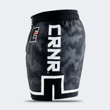 Left side profile of Combat Corner CRNR ISO black fight shorts. Features a large, vertical white "CRNR" logo running down the thigh, framed above a distinct white geometric border. A black side slit cut-out splits the lower hemline, and a small square orange and white logo is visible near the front hip.