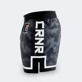 Left side profile of Combat Corner CRNR ISO black fight shorts. Features a large, vertical white "CRNR" logo running down the thigh, framed above a distinct white geometric border. A black side slit cut-out splits the lower hemline, and a small square orange and white logo is visible near the front hip.
