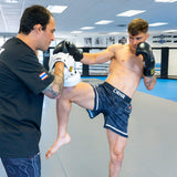 Shirtless fighter wearing Combat Corner CRNR ISO black fight shorts and black boxing gloves, throwing a knee strike into white focus mitts held by a coach. The action shot showcases the stretch and mobility of the shorts during intense training on grey gym mats.