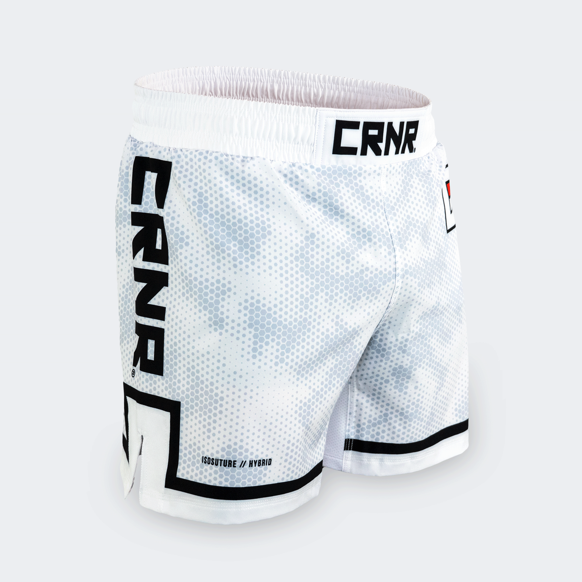 A pair of white CRNR ISO athletic fight shorts shown from a front-right three-quarter angle against a light gray background. The shorts feature an elastic waistband with "CRNR" printed in bold black letters on the front center. The fabric has a subtle white and gray hexagonal camo pattern. The right leg displays a large, vertical black "CRNR" logo, outlined at the bottom by a thick black geometric line wrapping around the side slit. The lower front right leg features the small text "ISOSUTURE // HYBRID" in 