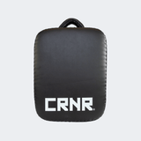 Back view of the HMITV Low Kick Pad Lite, a large, black leather martial arts low kick shield pad by Combat Corner against a light grey background, featuring a white block "CRNR" logo on the striking surface.