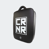 Front left angle view of the HMITV Low Kick Pad Lite, a large, black leather martial arts low kick shield pad by Combat Corner against a light grey background, featuring a white block "CRNR" logo on the striking surface.