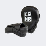 A pair of professional black HMITV Curved Punch Mitts by Combat Corner against a solid light grey background, showcasing the curved hand compartment and front striking surface.