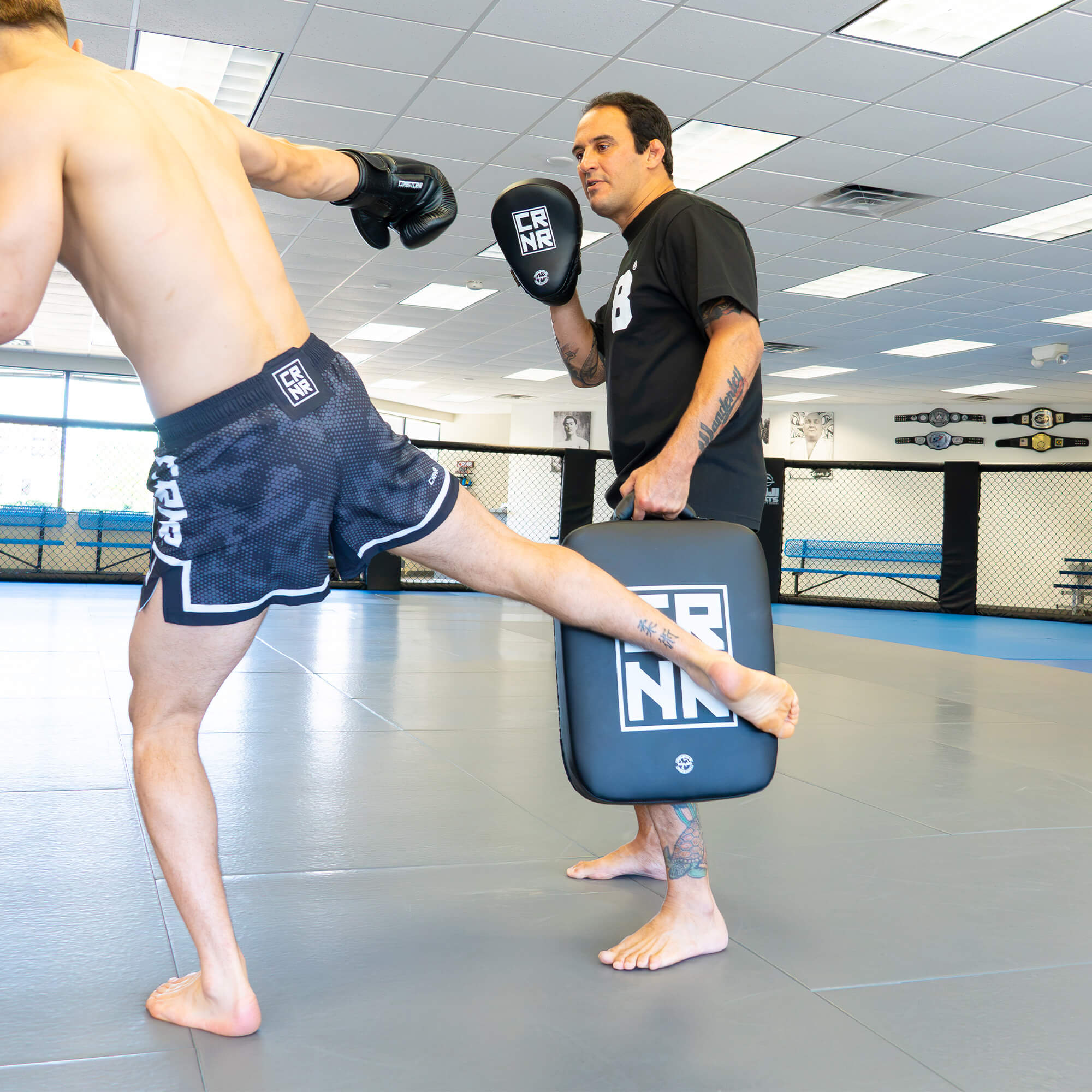 An action photograph of a pro MMA fighter executing a low kick against a the HMITV Low Kick Pad Lite held by a trainer, featuring professional training gear by Combat Corner.