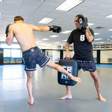 An action photograph of a pro MMA fighter executing a low kick against a the HMITV Low Kick Pad Lite held by a trainer, featuring professional training gear by Combat Corner.