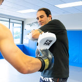 An action shot of a boxing trainer catching a close-range punch with the white HMITV Curved Punch Mitts, as a fighter strikes wearing black Combat Corner boxing gloves.