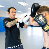 An action shot of a boxing trainer catching a close-range punch with the white HMITV Curved Punch Mitts, as a fighter strikes wearing black Combat Corner boxing gloves.