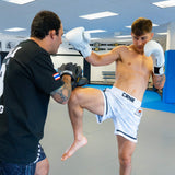 An athlete wearing white Combat Corner hybrid fight shorts and white boxing gloves throws a knee strike during a training session on grey gym mats. A coach wearing black Combat Corner focus mitts and a black t-shirt blocks the strike.